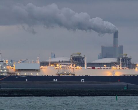 The VIVIT ARABIA LNG carrier docked at Europoort in Rotterdam, Netherlands, with industrial smokestacks and a wind turbine visible in the background. Photo: Wikimedia Commons / CC BY-SA 2.0