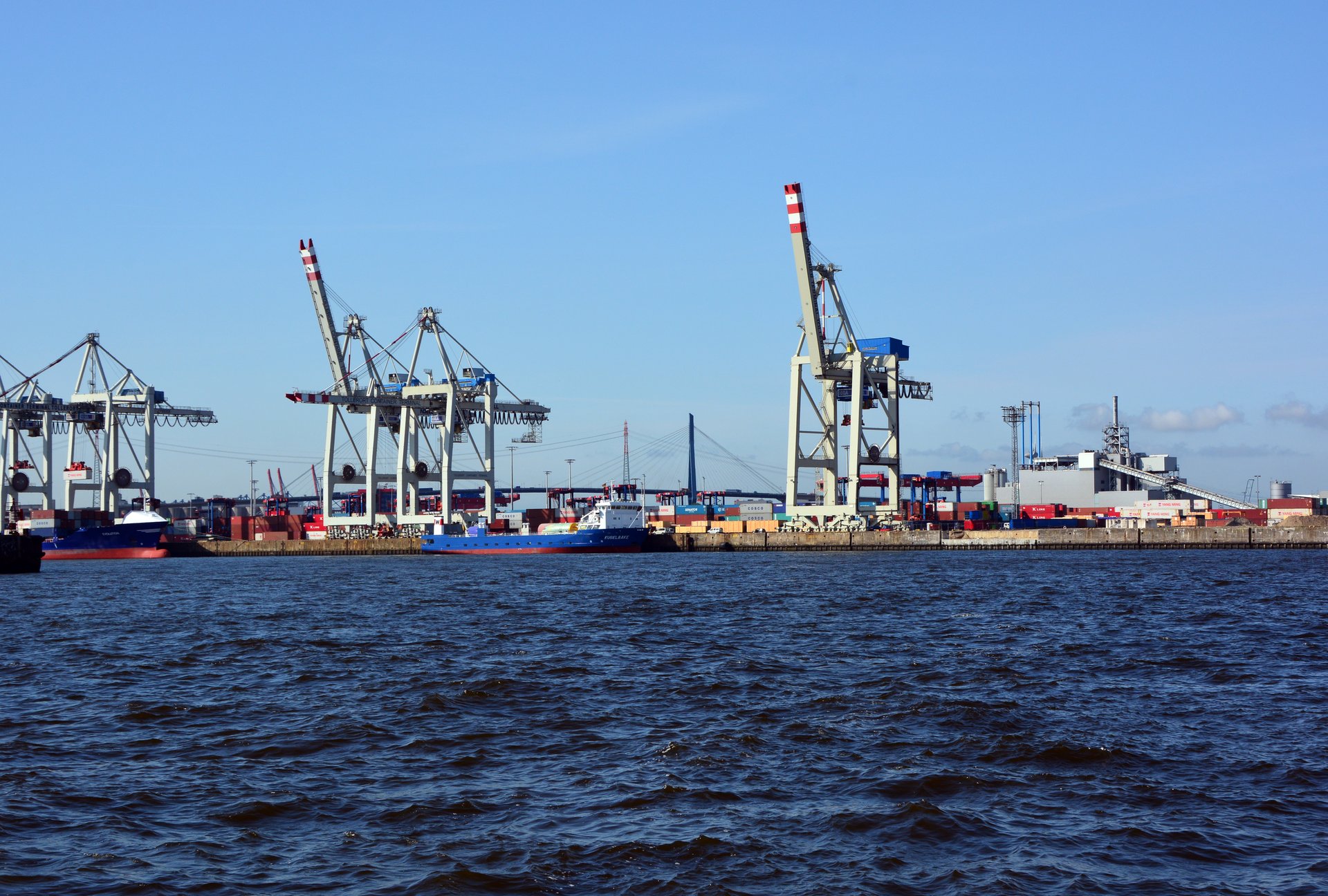 Container terminal with gantry cranes and cargo vessels, representing the global port infrastructure now under strain from the Hormuz shipping crisis. Photo: Wikimedia Commons / CC BY-SA 3.0
