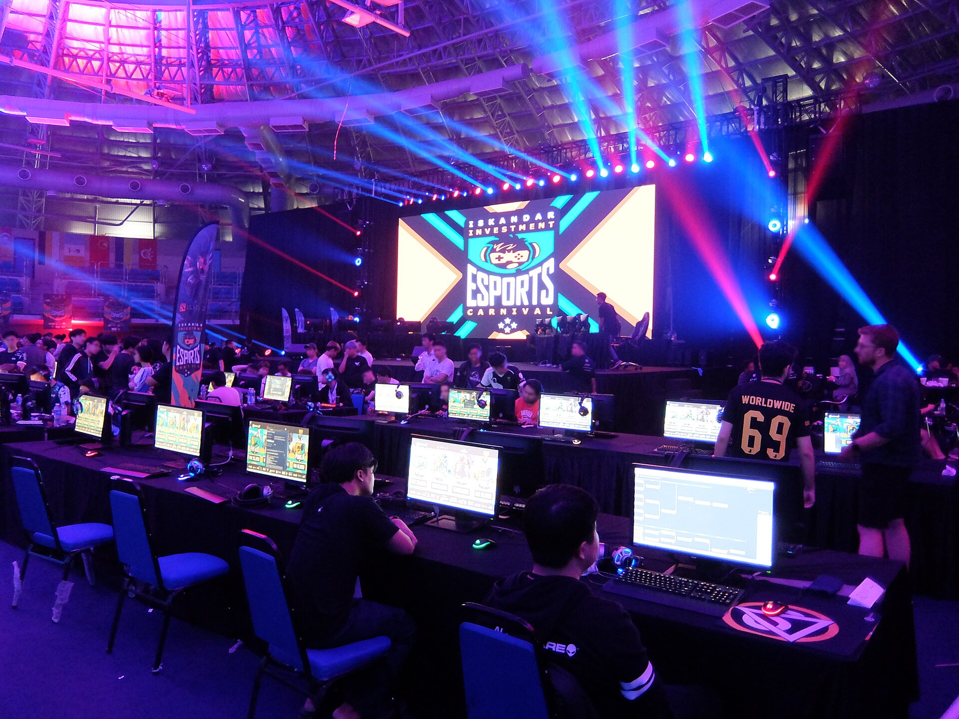 Esports tournament arena with competitors at gaming stations under stage lighting, representing Saudi Arabia billion-dollar gaming investment. Photo: Wikimedia Commons / CC BY-SA 4.0
