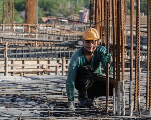 A migrant construction worker crouches among steel reinforcement bars on a Gulf building site, wearing a yellow hard hat and work gloves