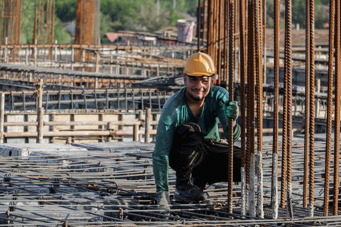 A migrant construction worker crouches among steel reinforcement bars on a Gulf building site, wearing a yellow hard hat and work gloves
