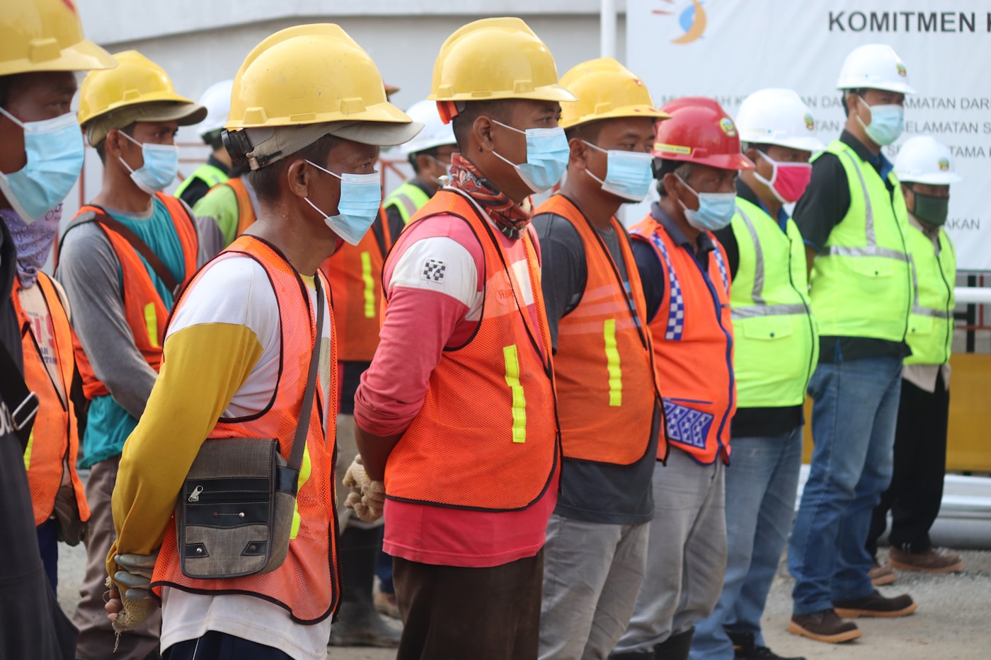Migrant construction workers wearing orange safety vests and yellow hard hats line up at a building site in the Gulf region