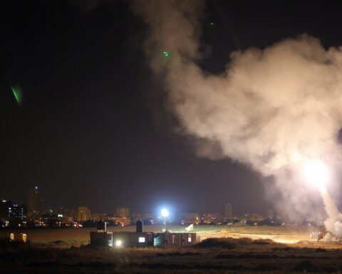 Iron Dome missile defense system intercepting rockets over an Israeli city at night, with smoke trails visible against the dark sky. Photo: Wikimedia Commons / CC BY 2.0
