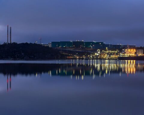 Oil refinery illuminated at dusk with industrial towers and storage tanks reflected in water, representing the global energy crisis triggered by the 2026 Iran war. Photo: Wikimedia Commons / CC BY-SA 4.0