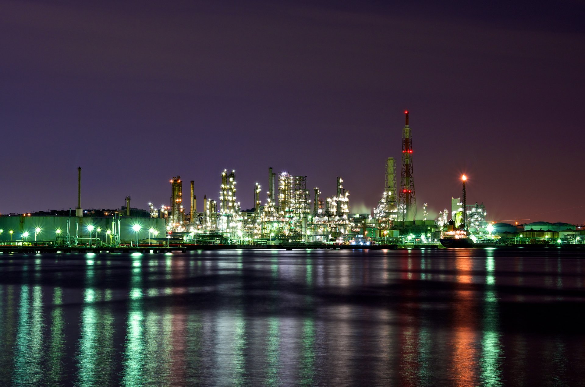 Oil refinery illuminated at night with distillation columns and industrial towers reflected in waterfront, representing the downstream petroleum infrastructure affected by Gulf war risk premiums