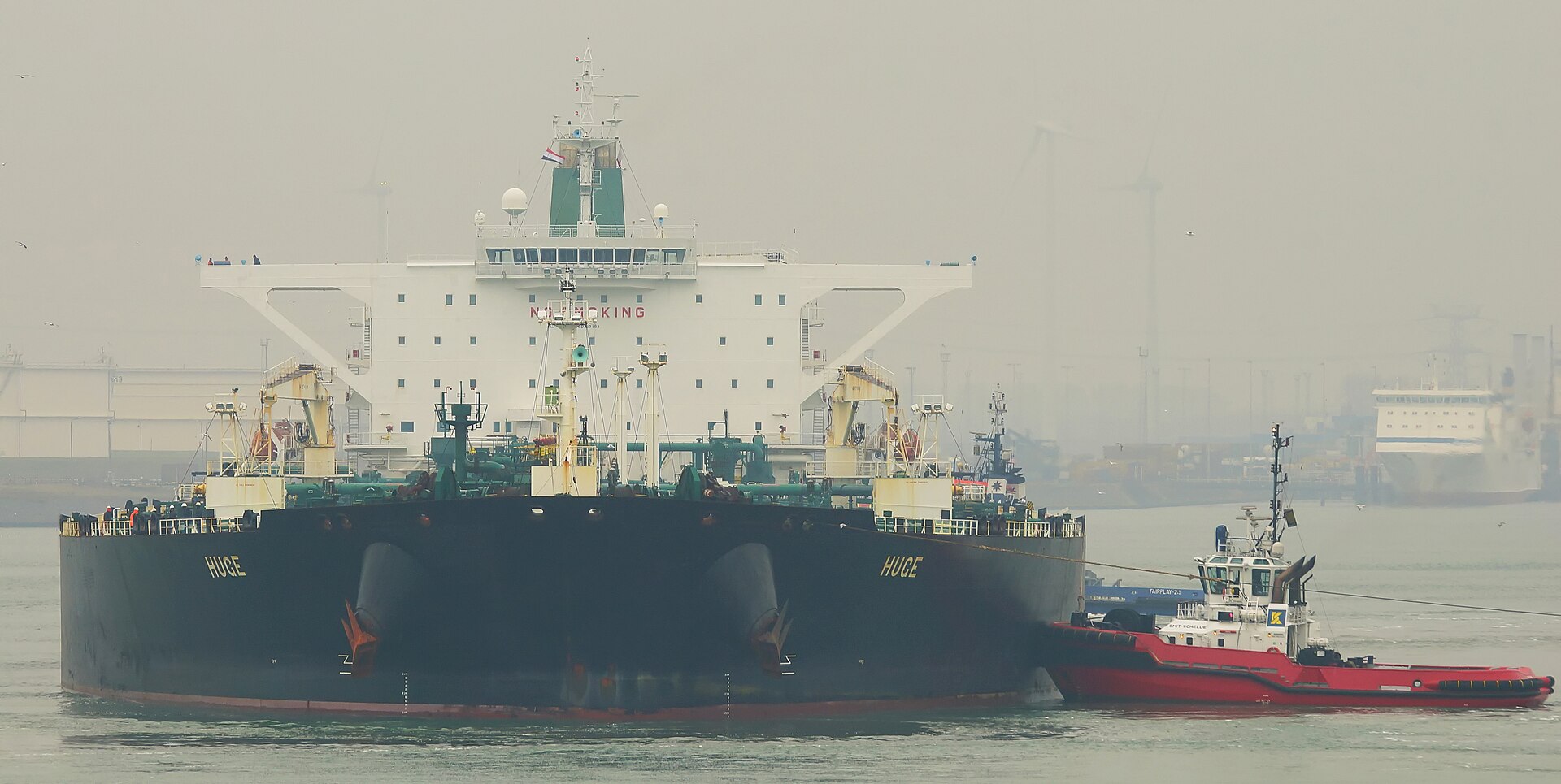 A large crude oil tanker being guided by tugboats in port, representing the stranded shipping fleet unable to transit the Strait of Hormuz during the 2026 Iran war