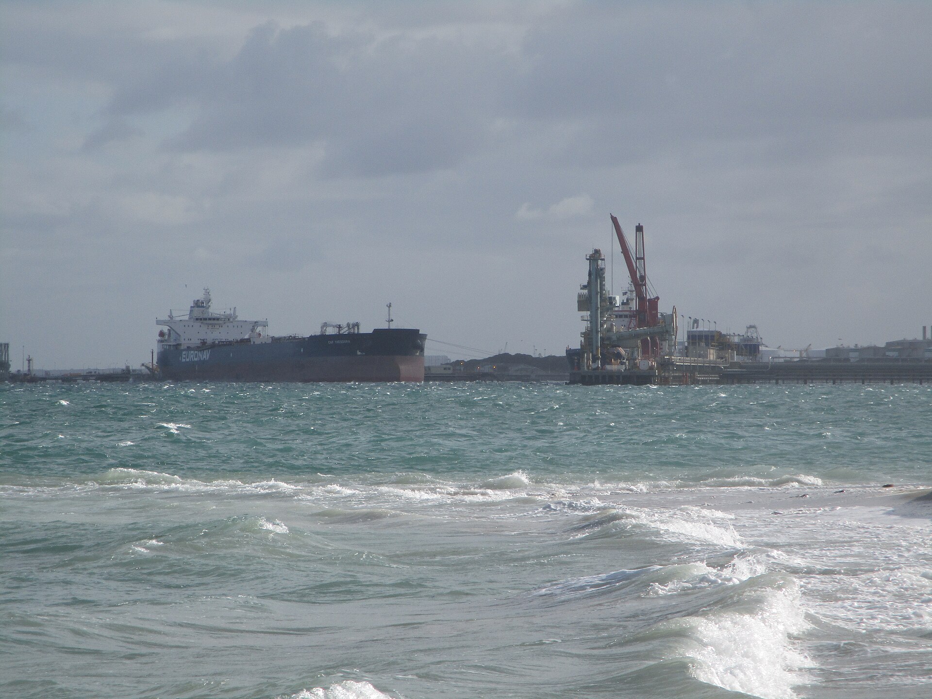 Oil tanker docked at a refinery port terminal with loading cranes, representing the global oil export infrastructure disrupted by the Strait of Hormuz blockade. Photo: Wikimedia Commons / CC BY-SA 4.0