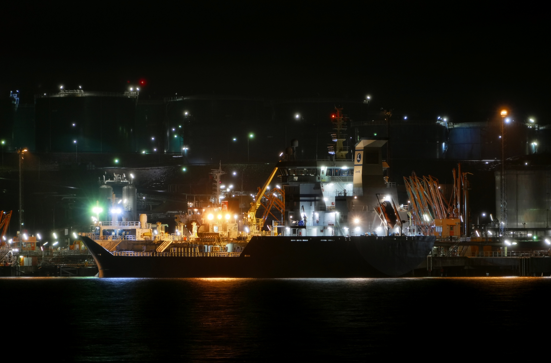 Oil tanker loading at a petroleum product terminal at night with storage tanks and port infrastructure illuminated