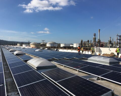 Solar photovoltaic panels installed on a rooftop adjacent to oil refinery infrastructure, symbolizing the global energy transition from fossil fuels to renewable sources. Photo: US Department of Energy / Public Domain