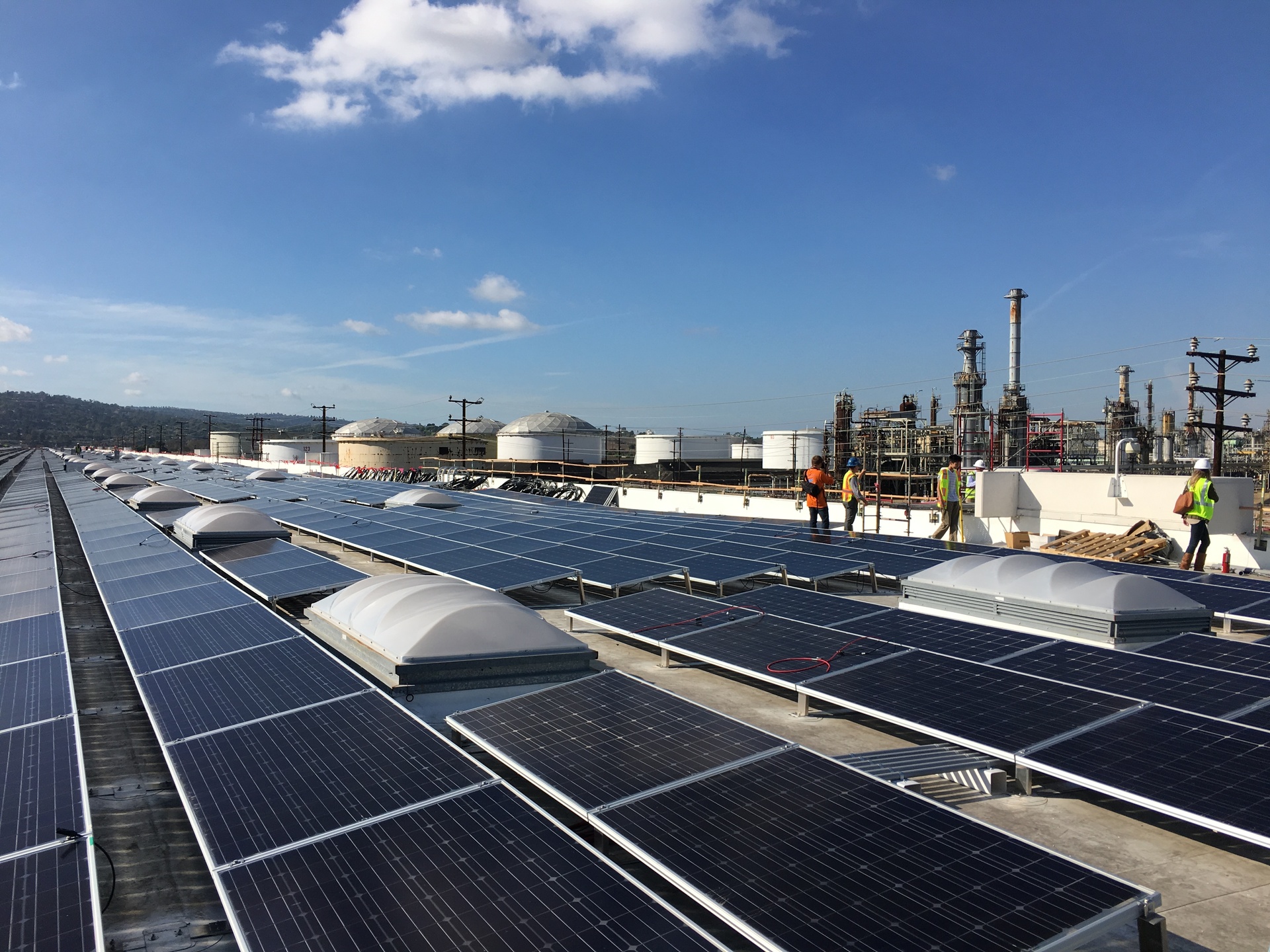 Solar photovoltaic panels installed on a rooftop adjacent to oil refinery infrastructure, symbolizing the global energy transition from fossil fuels to renewable sources. Photo: US Department of Energy / Public Domain