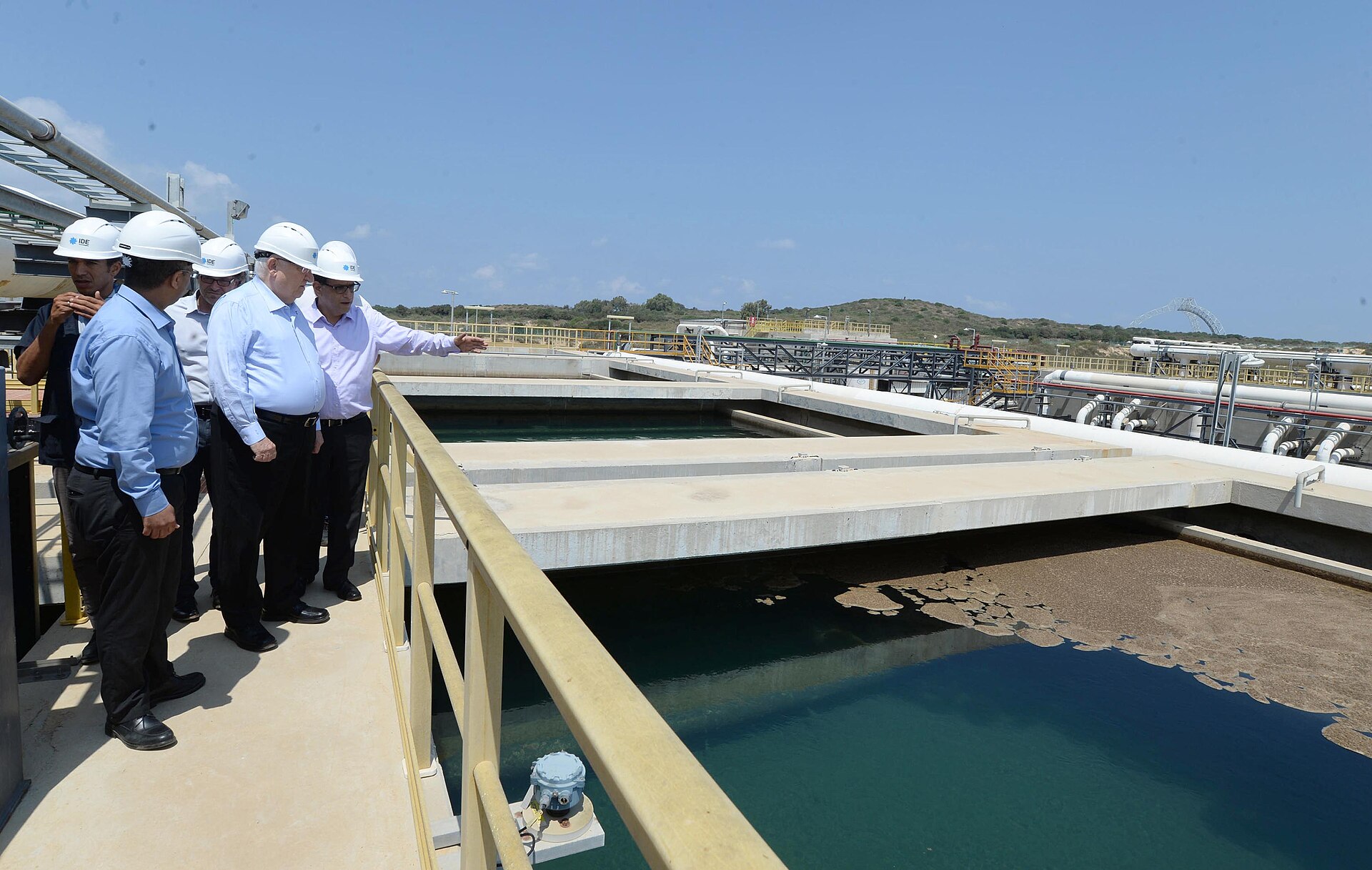 Officials in hard hats touring a desalination plant with open water treatment basins and industrial infrastructure along the coast