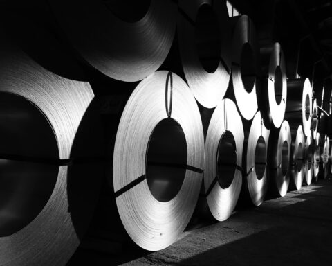 Rolls of steel and metal coils stacked in an industrial warehouse, representing the global metals supply chain disrupted by the Strait of Hormuz closure
