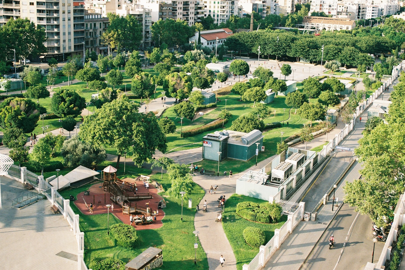 An aerial view of a modern urban park with green spaces and tree-lined pathways in a dense city environment