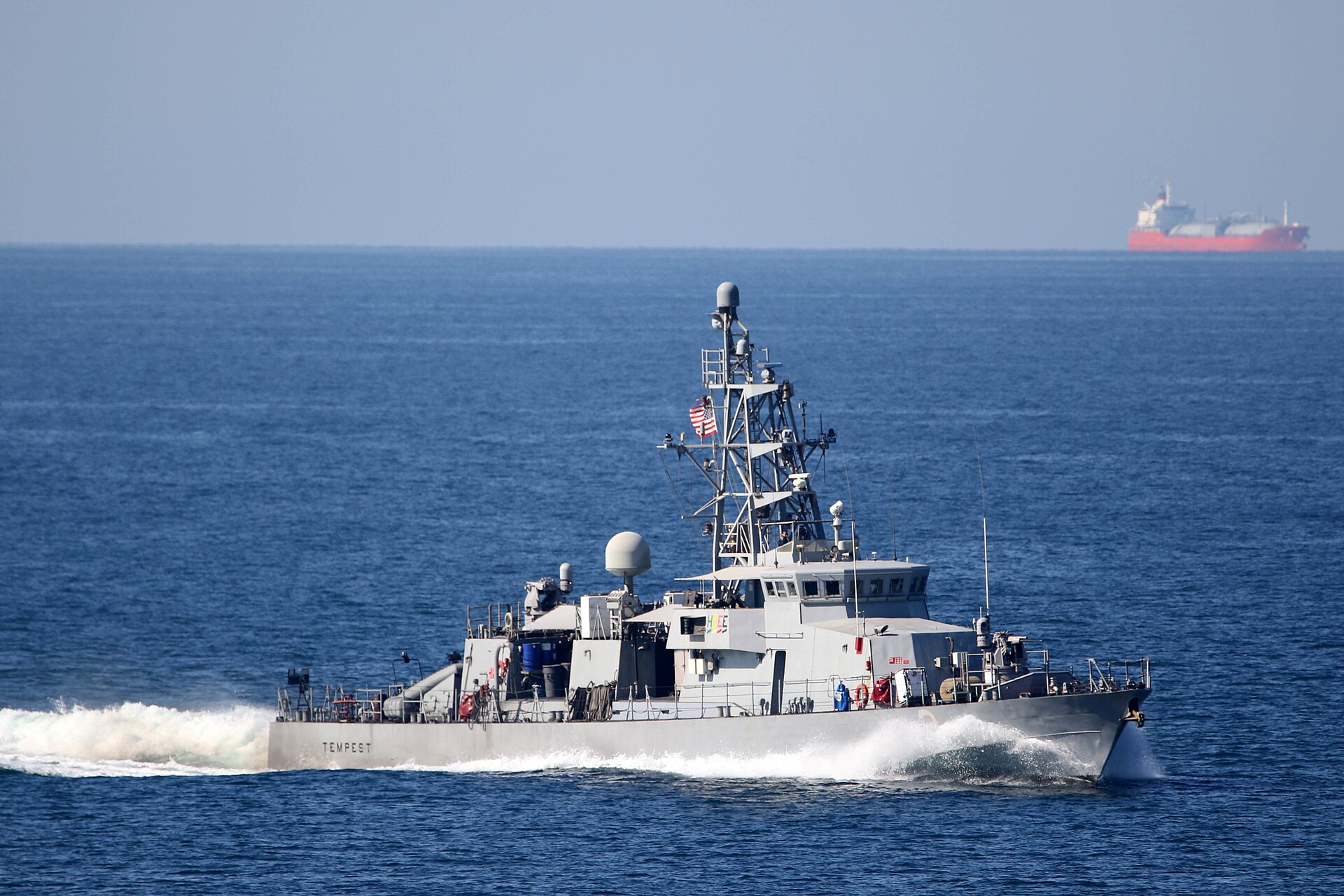 USS Tempest patrol craft transits the Strait of Hormuz with an oil tanker visible in the background. The strait remains nearly impassable to commercial shipping amid the Iran war. Photo: US Navy / Public Domain