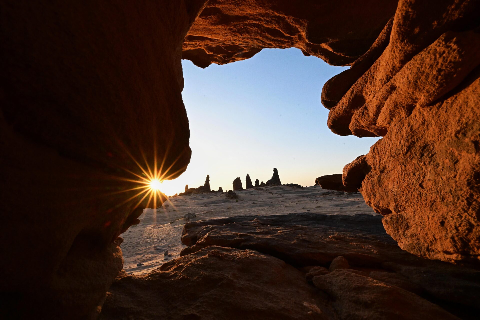 Sunset through a natural sandstone cave arch in AlUla, Saudi Arabia, with desert rock formations silhouetted against the sky