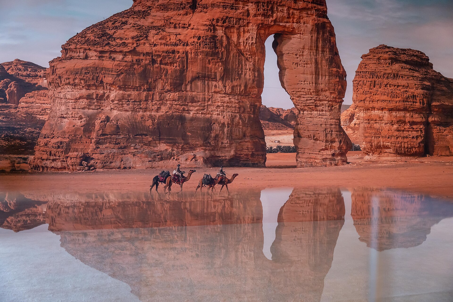 Elephant Rock natural sandstone formation in AlUla at sunset with camel riders and reflections in water