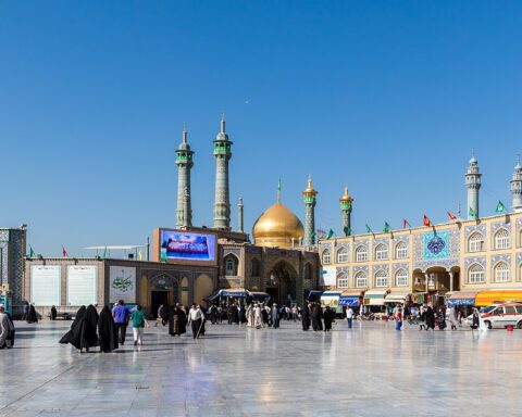 The Fatima Masumeh shrine in Qom, Iran — exterior panoramic view showing the golden dome and minarets, with pilgrims in the courtyard