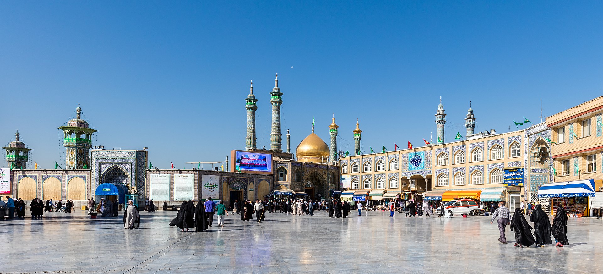 The Fatima Masumeh shrine in Qom, Iran — exterior panoramic view showing the golden dome and minarets, with pilgrims in the courtyard
