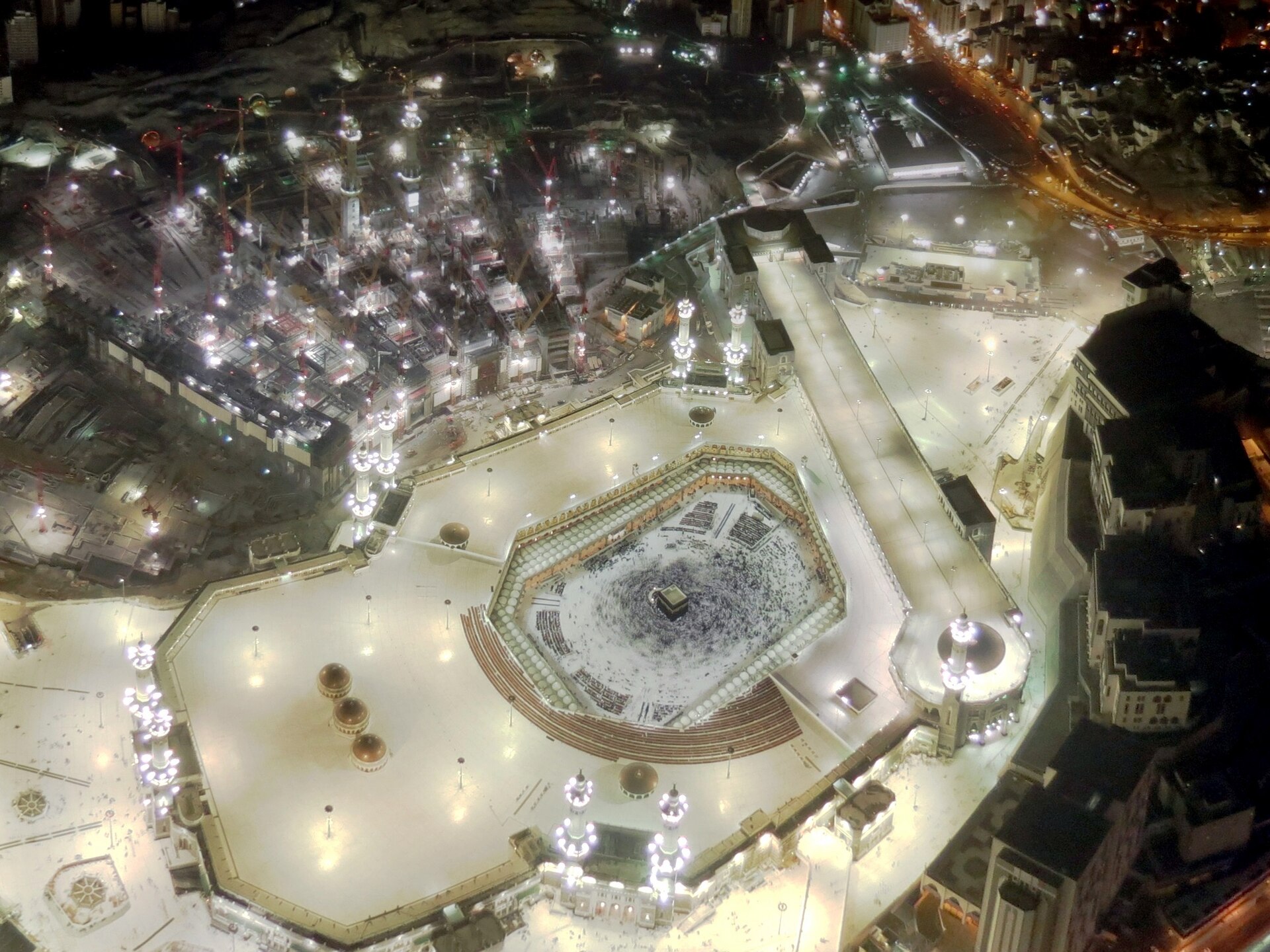 Aerial night view of Masjid al-Haram in Mecca showing the Kaaba surrounded by pilgrims performing tawaf, with mosque expansion construction cranes visible