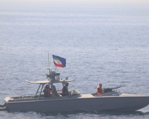 IRGC armed fast-attack gunboat with mounted machine gun patrolling the Strait of Hormuz, Iranian flag visible, crew in orange vests
