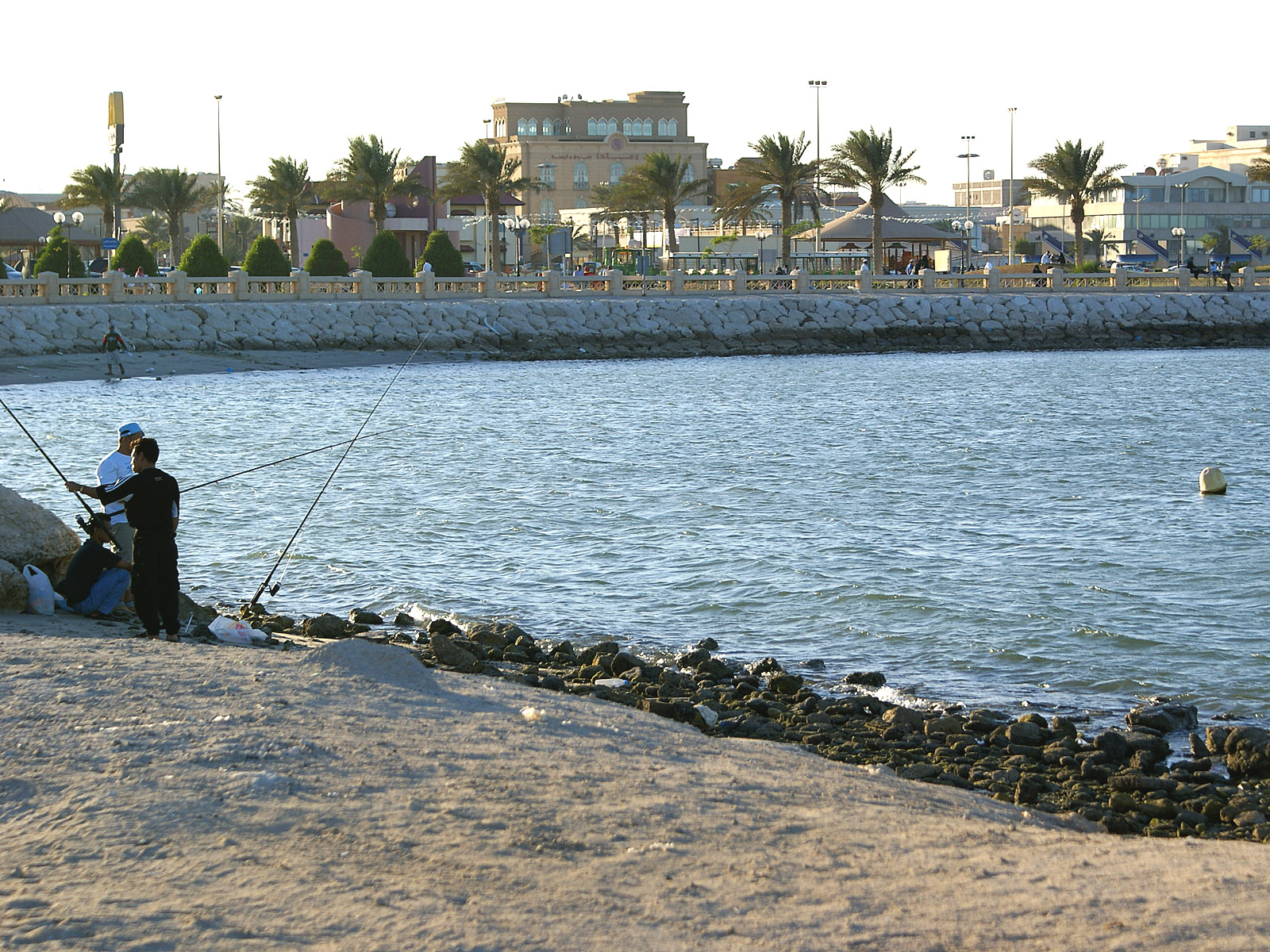 Fishermen casting lines along the Al Khobar Corniche at golden hour, with the palm-lined promenade and waterfront buildings visible in the background