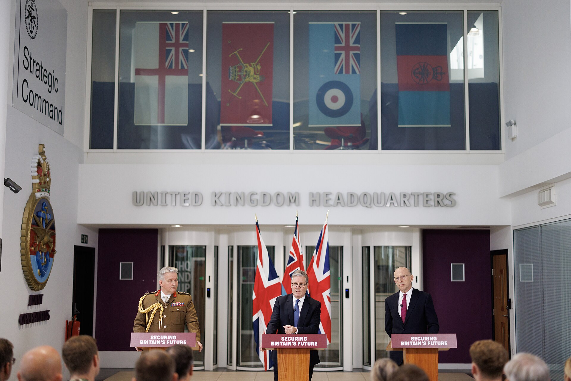 UK Strategic Command interior at Permanent Joint Headquarters Northwood where Keir Starmer addressed military staff below service flags of Royal Navy, Army, RAF, and Royal Marines