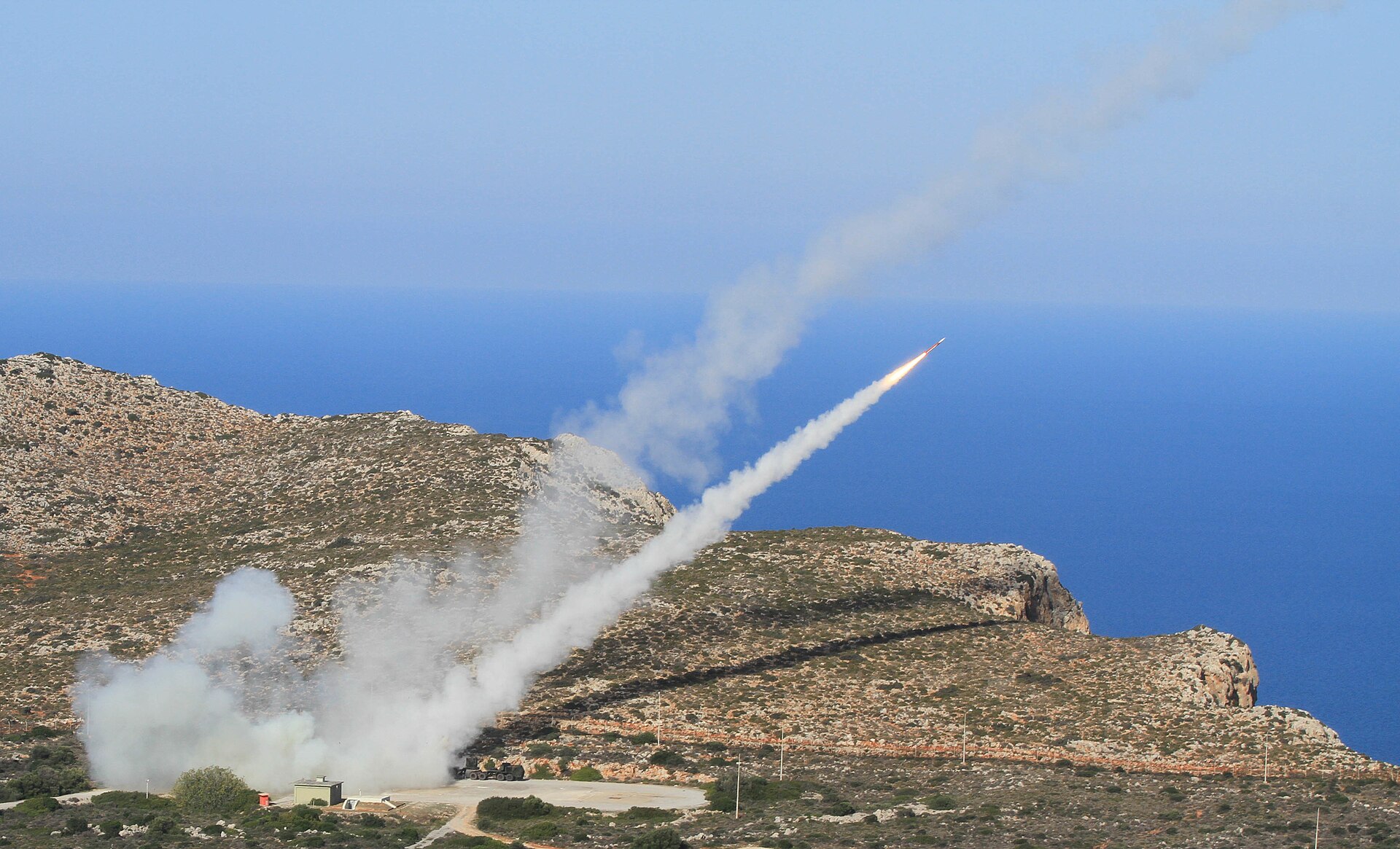 US Army Patriot missile system firing during Artemis Strike 2017 live-fire exercise, with missile contrail visible against blue sky over rocky Mediterranean terrain