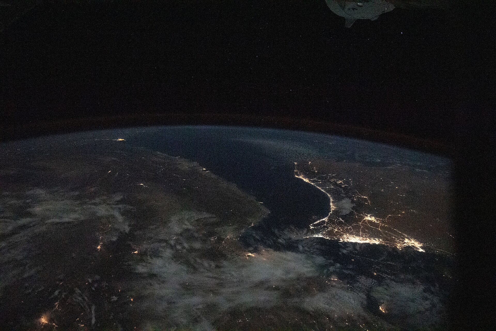 Persian Gulf and Gulf of Oman at night photographed from the International Space Station, Expedition 64 — the lit coastlines trace the Gulf states whose export revenues depend on Hormuz remaining open