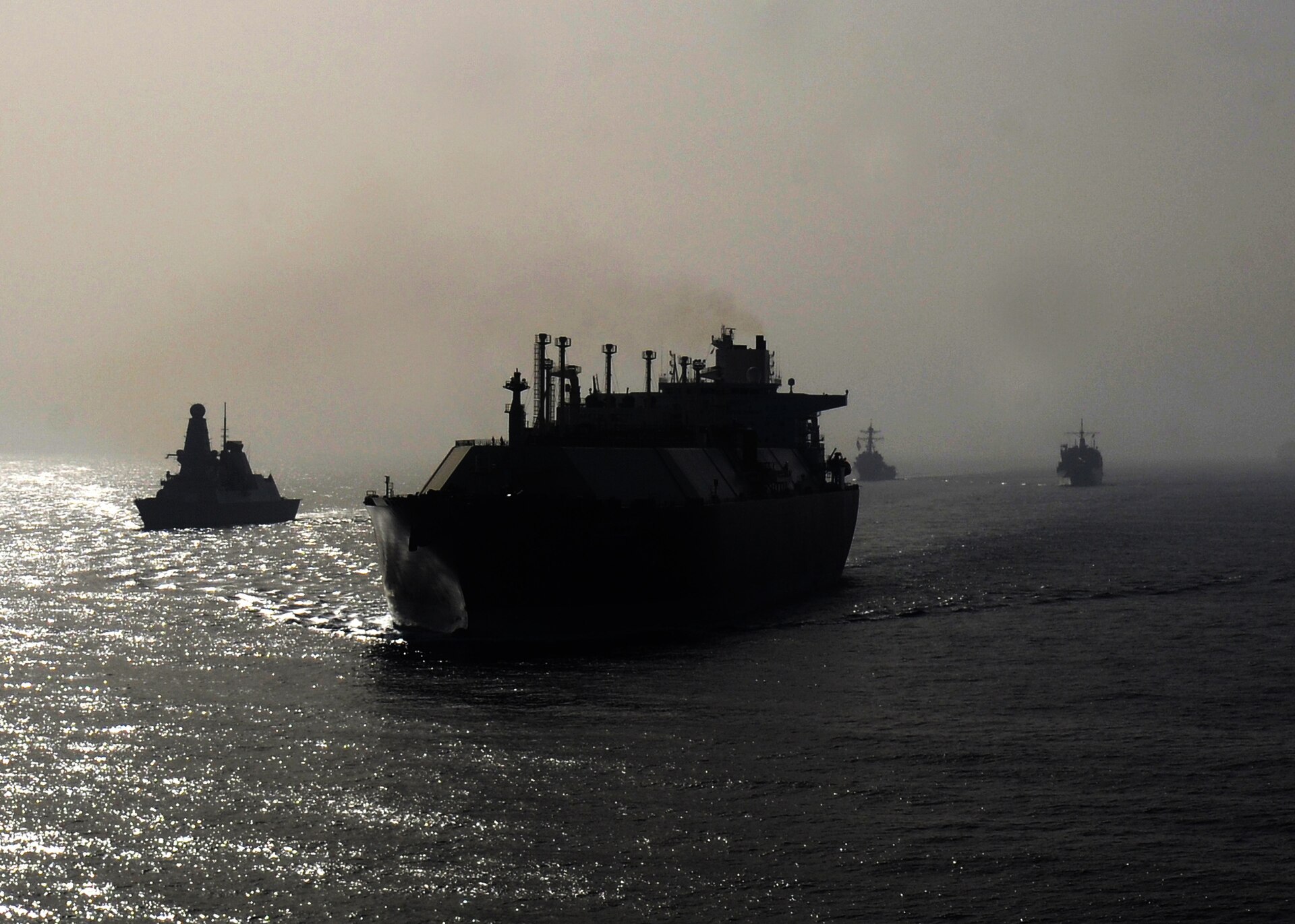 A large natural gas tanker silhouetted against the Persian Gulf haze, flanked by US Navy warships during Combined Task Force 521 convoy escort operations, 2013
