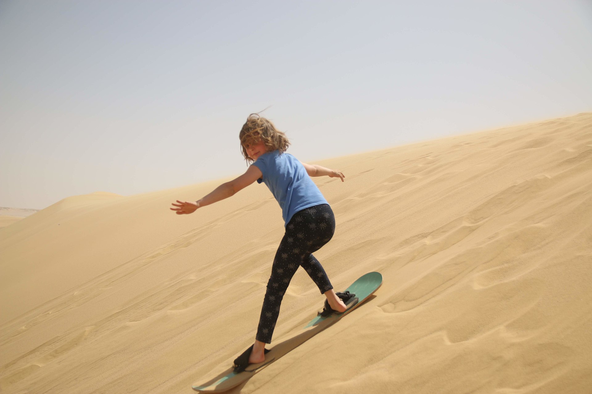 A young sandboarder sliding down a large sand dune, demonstrating the seated technique popular with beginners