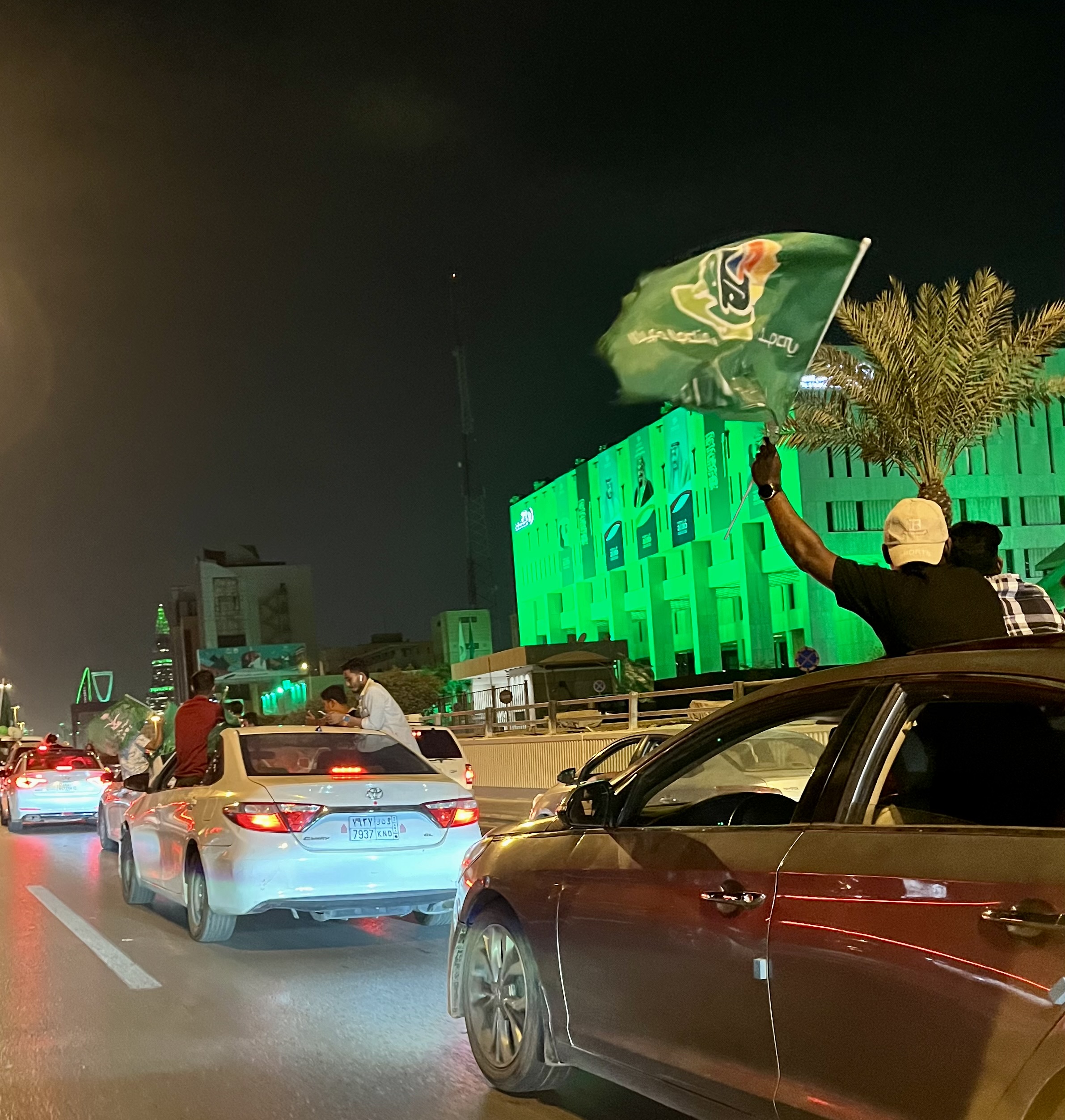 Cars waving Saudi flags with green-lit buildings during National Day street celebrations