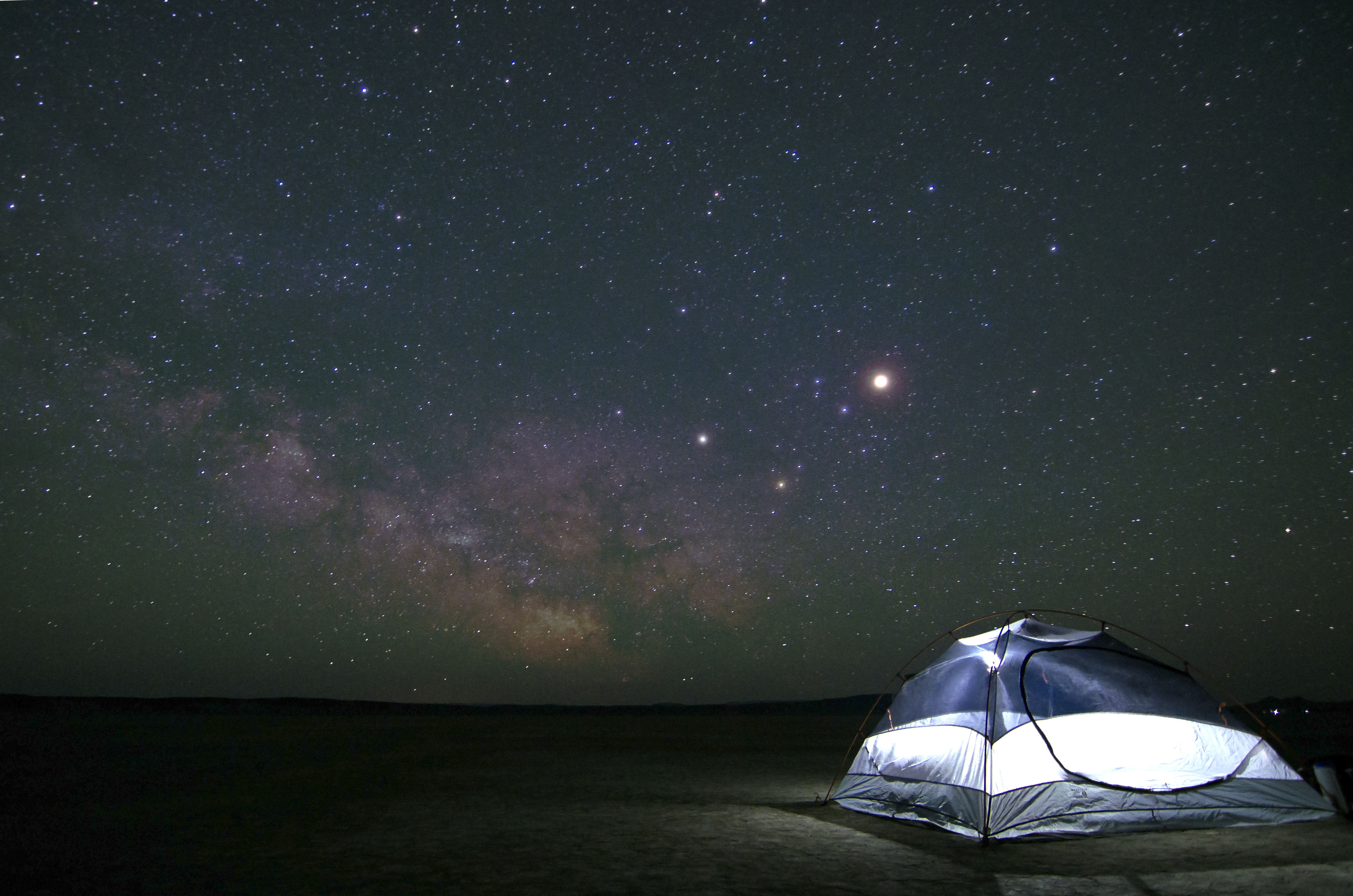 A glowing tent pitched under a vast night sky filled with the Milky Way in a flat desert landscape