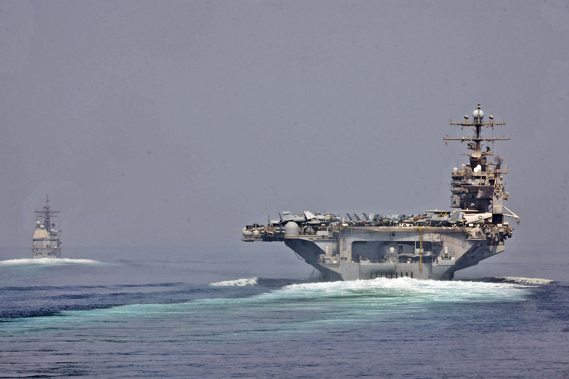 USS Abraham Lincoln carrier strike group transiting the Strait of Hormuz in 2012, accompanied by the guided-missile cruiser USS Cape St. George — the same transit route Iran now subjects to a $2 million per-vessel toll under IRGC control