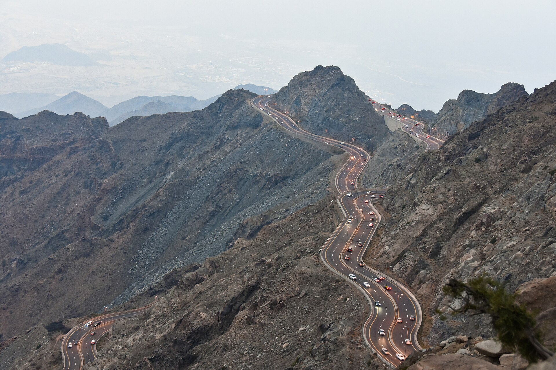 The winding Al Hada mountain road seen from above at dusk, with car headlights tracing the switchbacks through the Sarawat Mountains near Taif