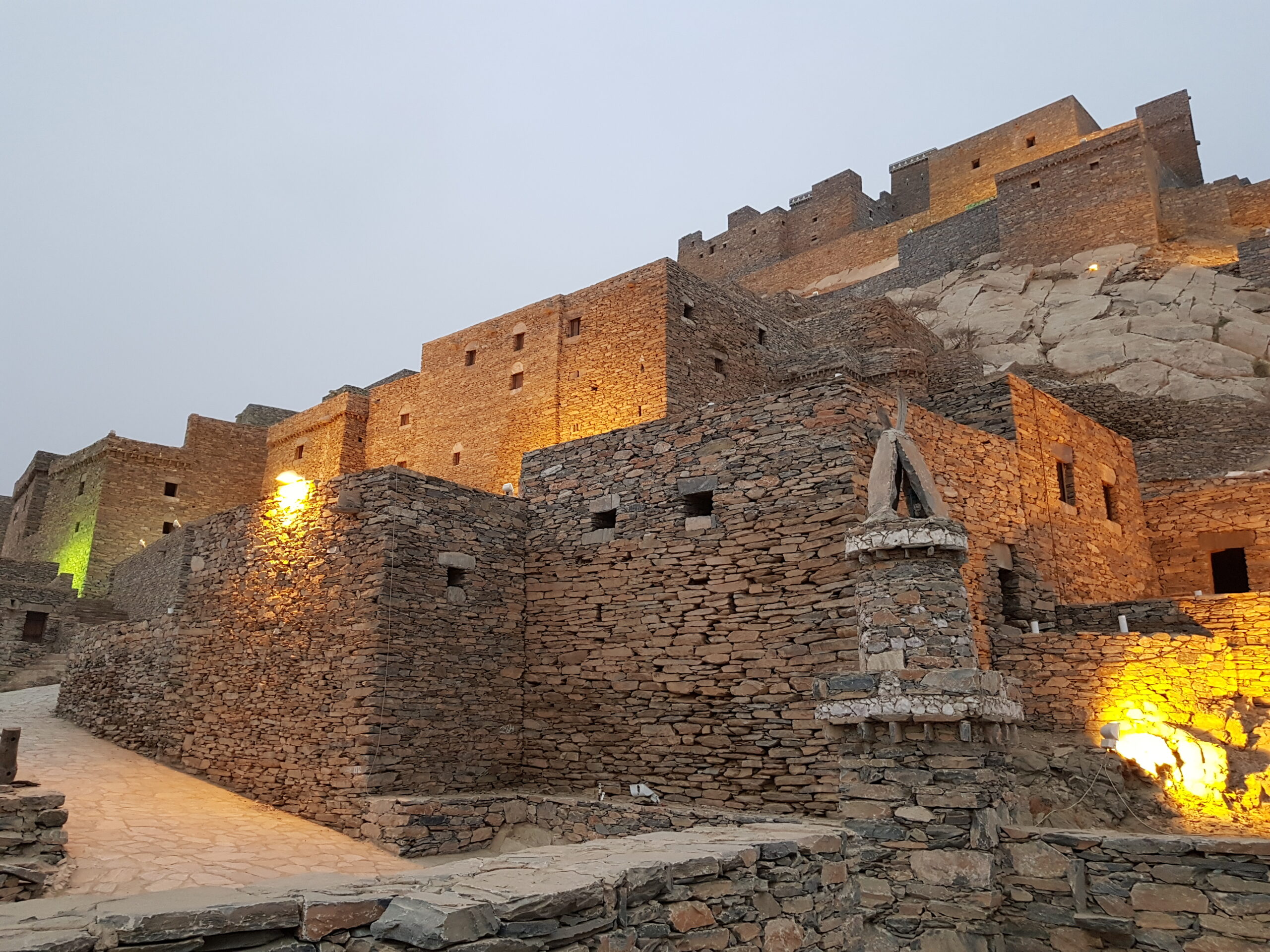 Thee Ain village stone buildings illuminated at dusk with warm golden lighting against twilight sky