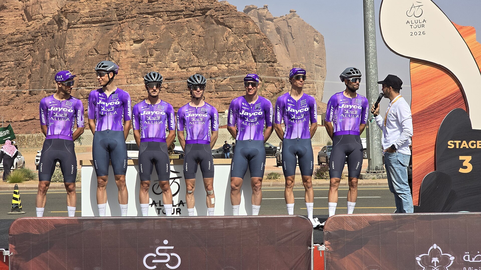 Professional cyclists from Team Jayco AlUla lined up at Stage 3 of the AlUla Tour 2026 with sandstone rock formations in the background