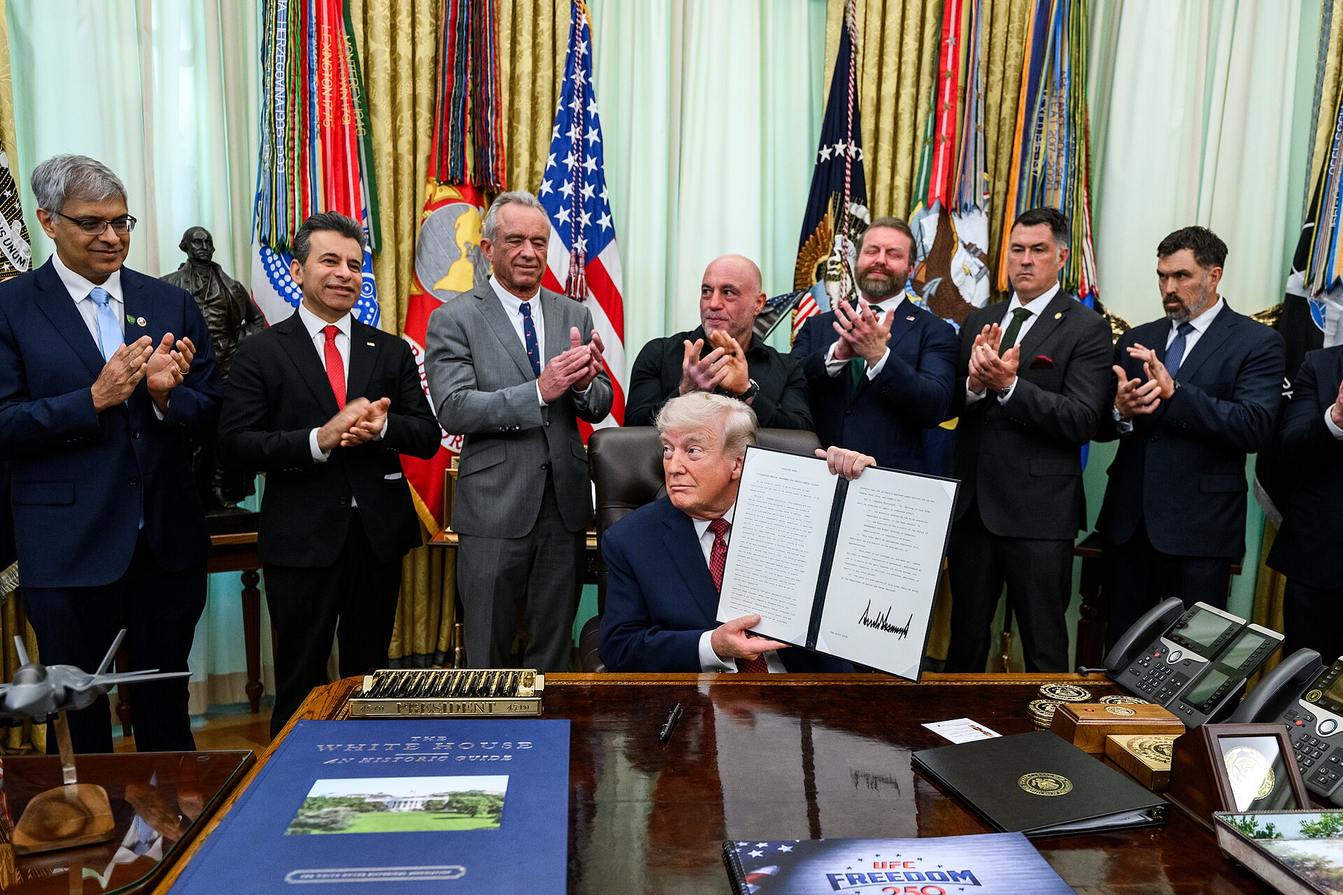 President Donald Trump in the Oval Office, April 2026, holding a signed executive order as advisers applaud — the same setting from which he declared on April 23 that he is in no rush to end the Iran crisis