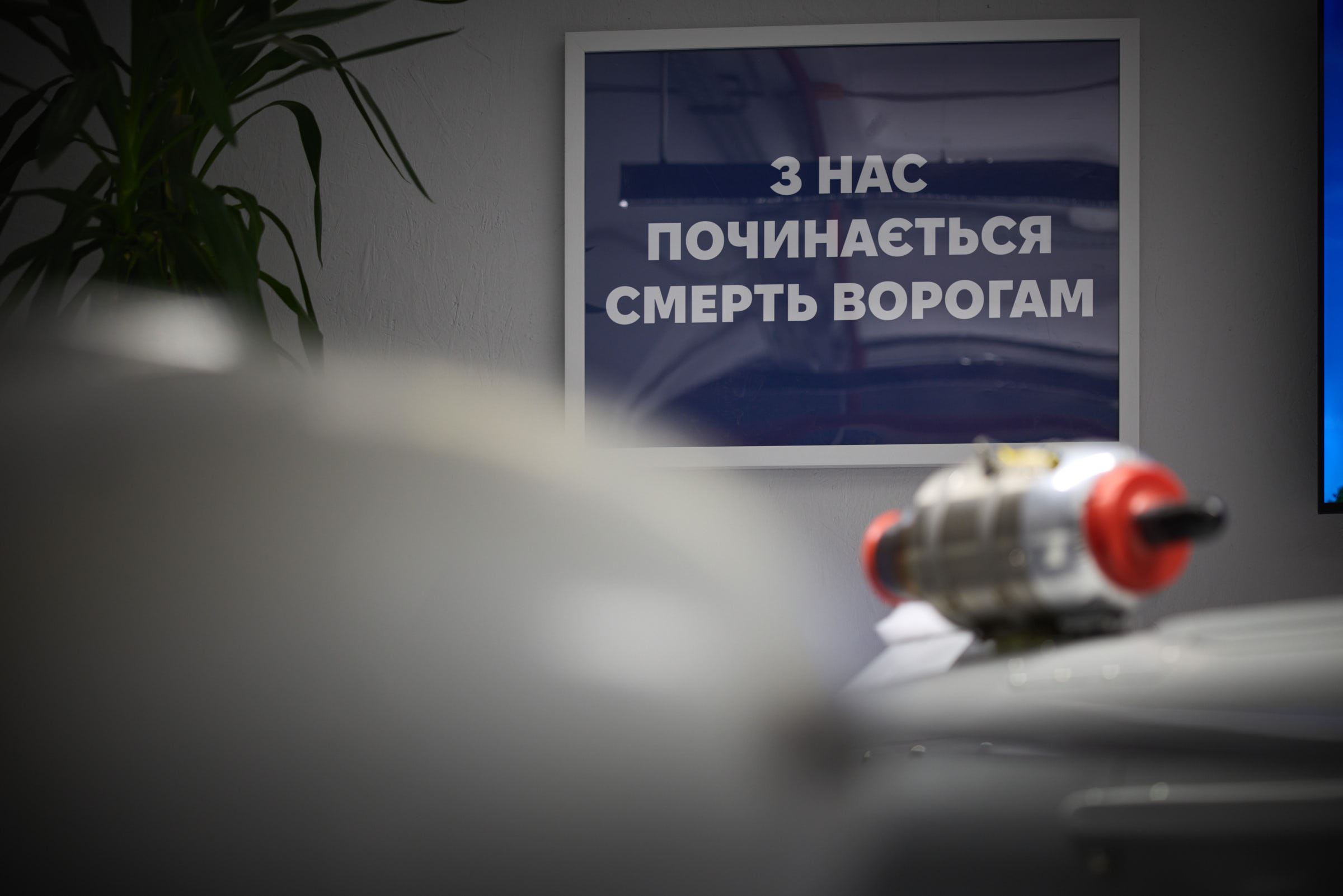 Inside a Ukrainian defense production facility, with drone and weapons hardware visible beneath a sign reading From us begins the death of enemies — the distributed, rapid-iteration manufacturing model Zelenskyy is proposing to export to Saudi Arabia