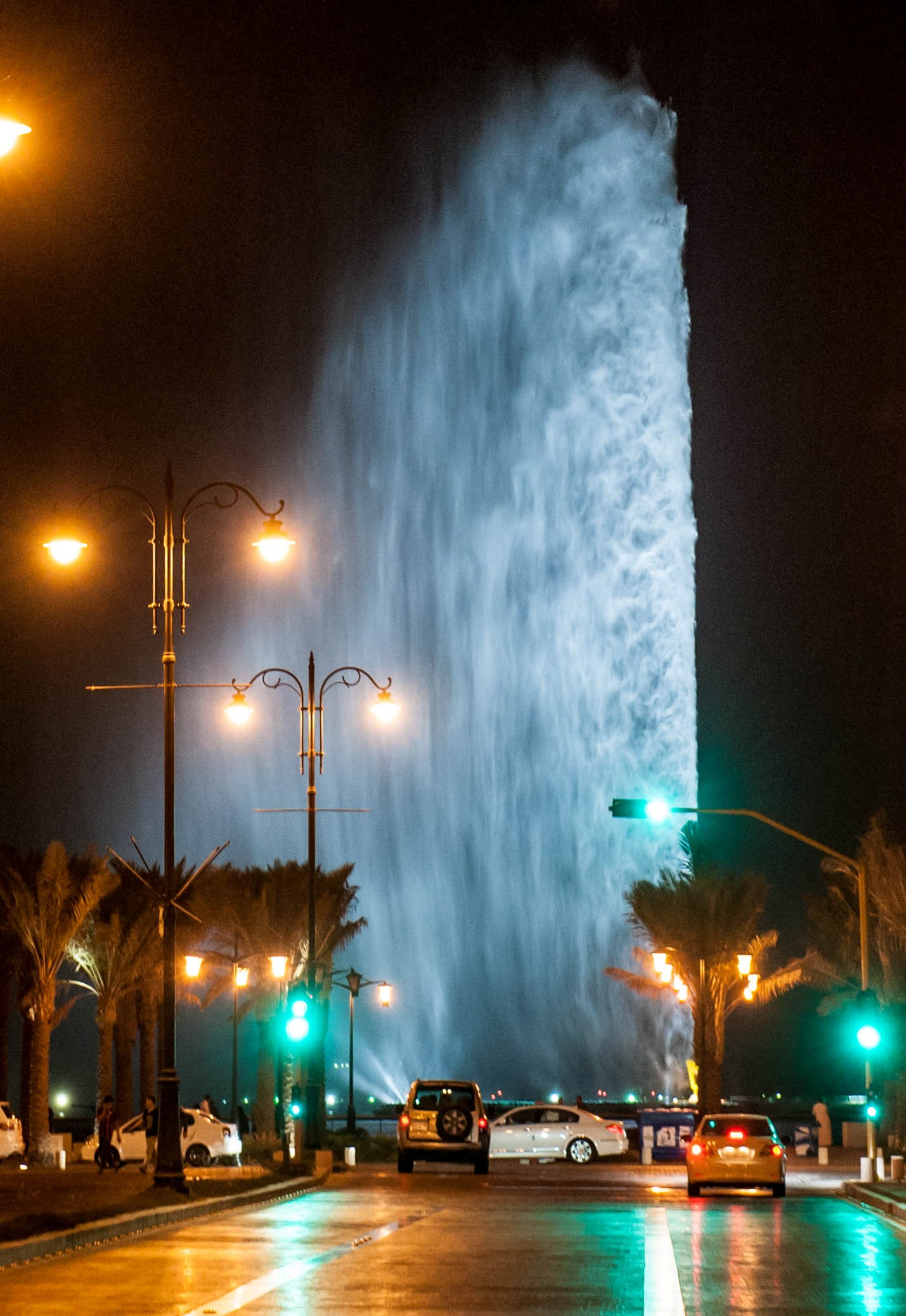 King Fahd Fountain in Jeddah illuminated at night, with the massive water jet soaring into the dark sky above palm-lined streets