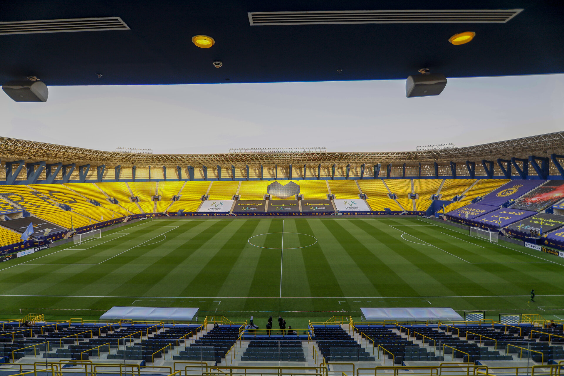 Interior view of Al-Awwal Park from pitch level showing the yellow and blue seating tiers and floodlights