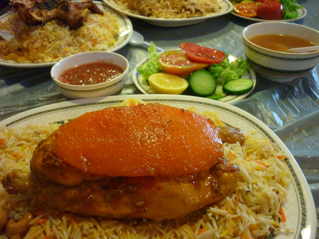 Plate of chicken mandi on saffron-tinted rice, with fresh tomato and cucumber salad, red chilli sauce and a bowl of broth