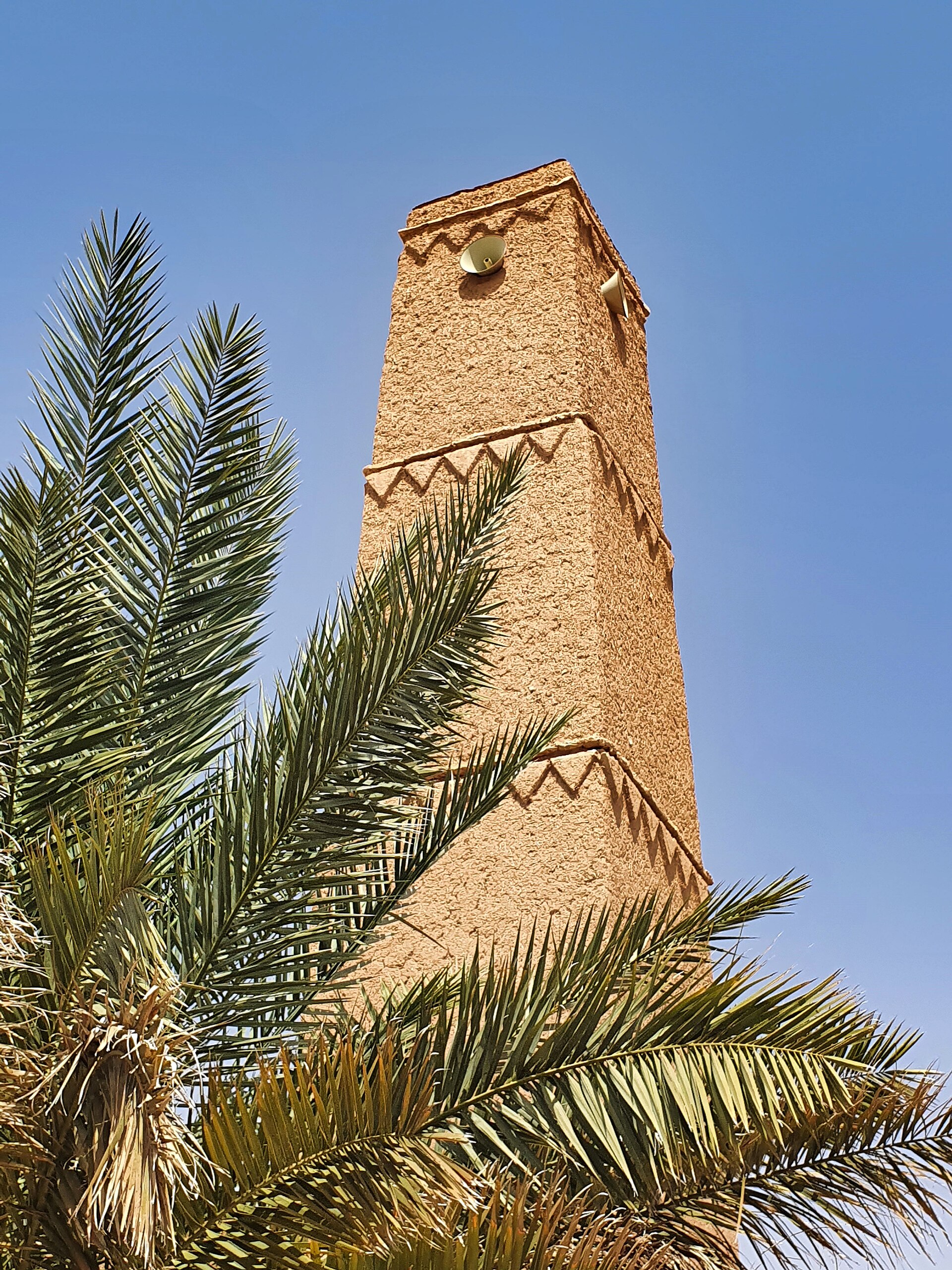 Traditional mud-brick minaret of a mosque in Shaqra, Saudi Arabia, framed by palm fronds
