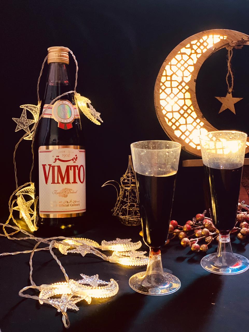 A bottle of Vimto fruit cordial with two glasses of the dark purple drink, decorated with Ramadan crescent moon and star lights