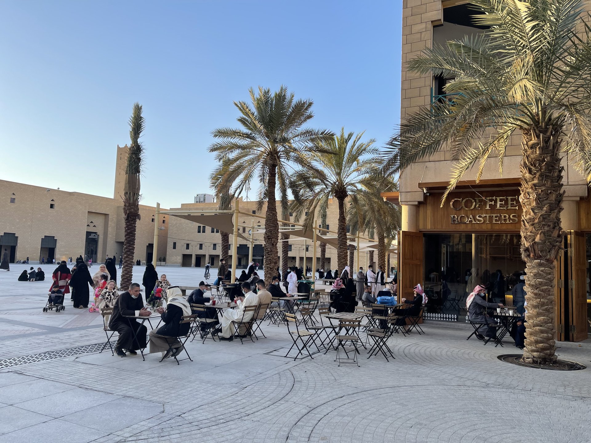 An outdoor coffee shop with patrons seated at tables under palm trees in Deera Square, Riyadh, Saudi Arabia
