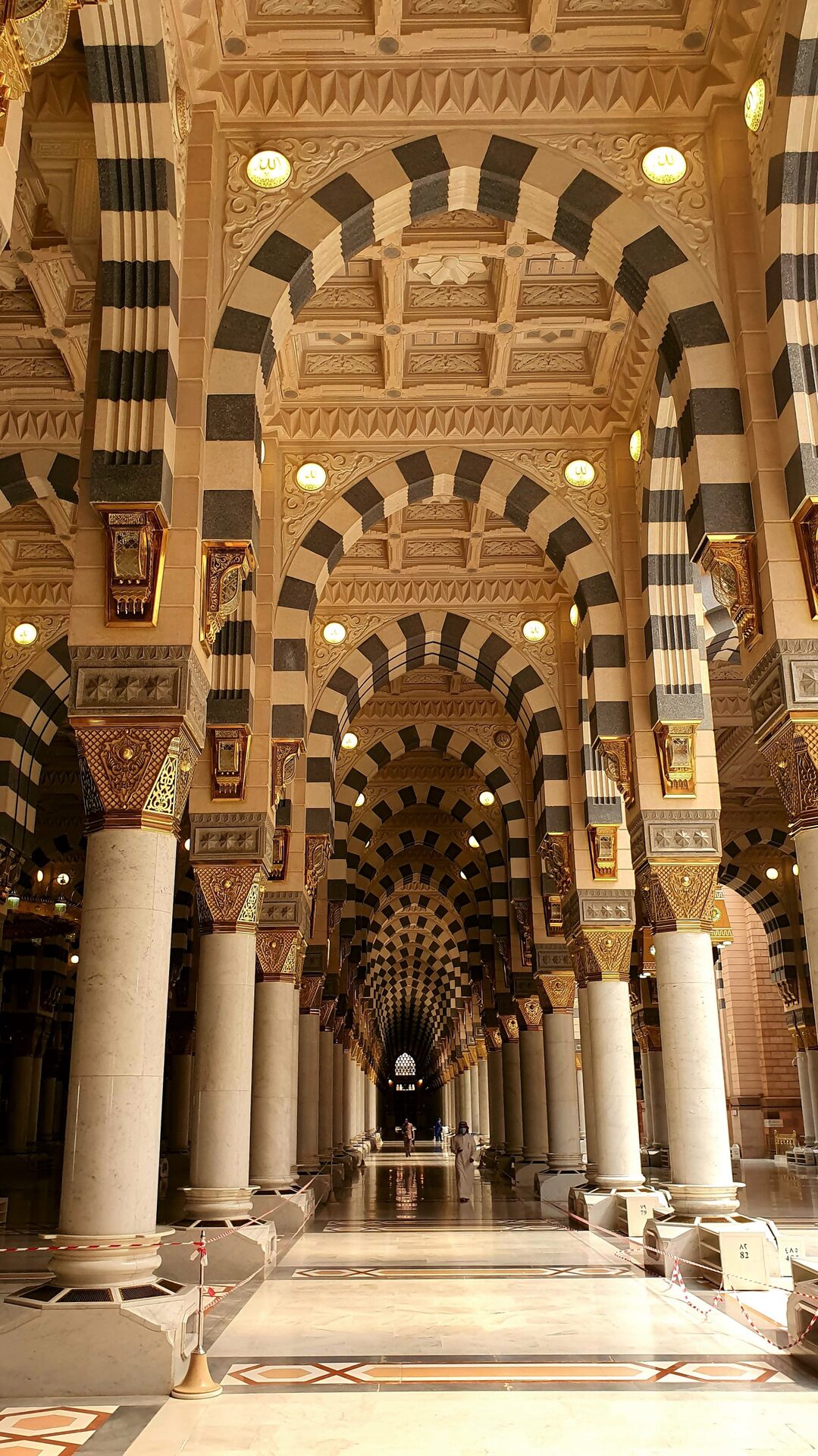 The ornate interior of the Prophet's Mosque in Madinah, Saudi Arabia, with striped arches and golden columns