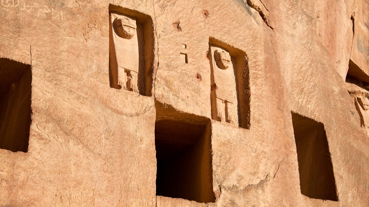 Rock-cut tombs carved into the red sandstone cliff face at Dadan, AlUla, featuring lion sculptures flanking burial niches