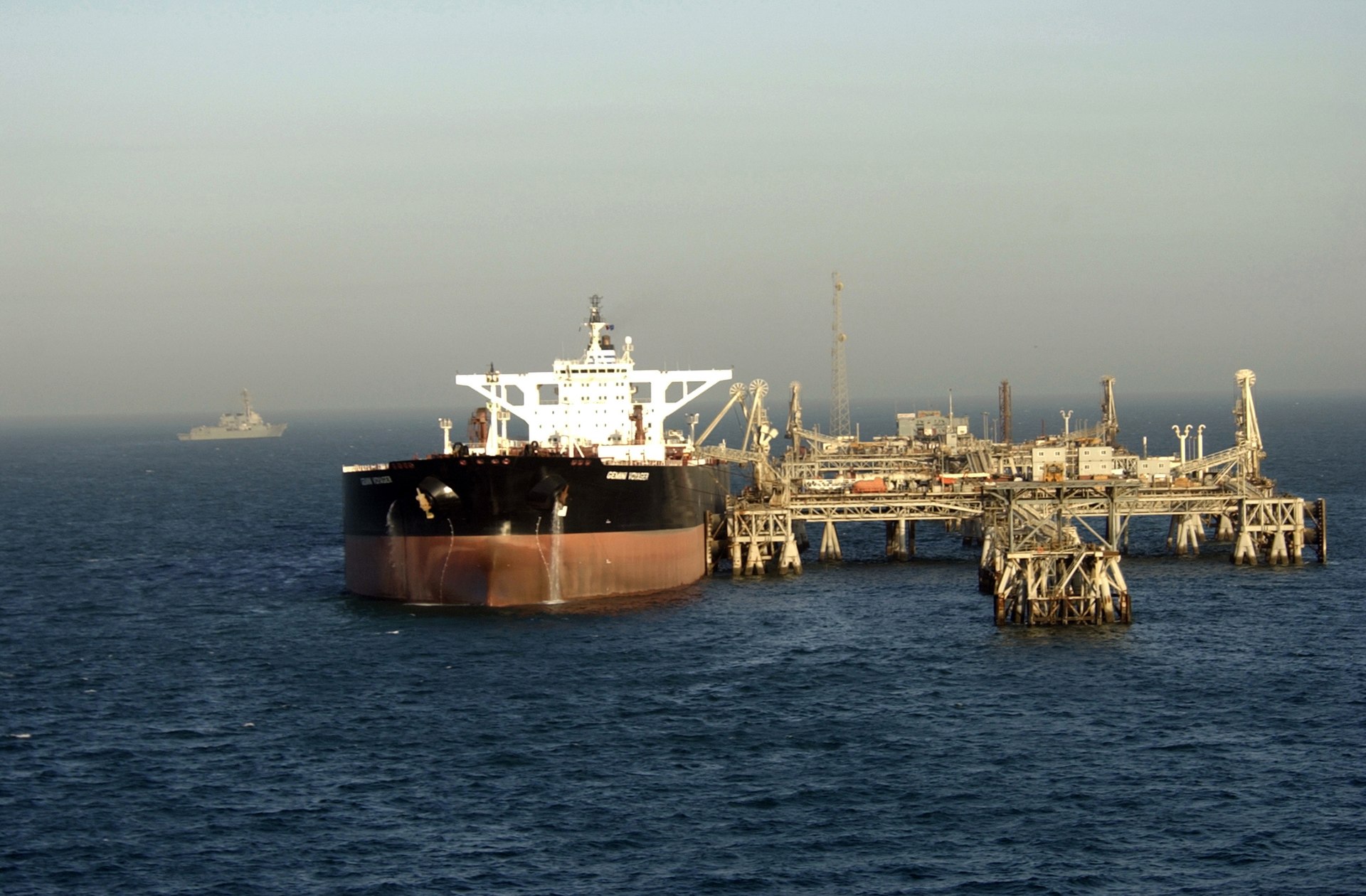 A VLCC supertanker takes on crude oil at the Al Basrah Oil Terminal in the Persian Gulf, with a US Navy guided missile destroyer visible on patrol in the background — the inverse of the current Hormuz situation, where IRGC rather than US forces are controlling tanker access