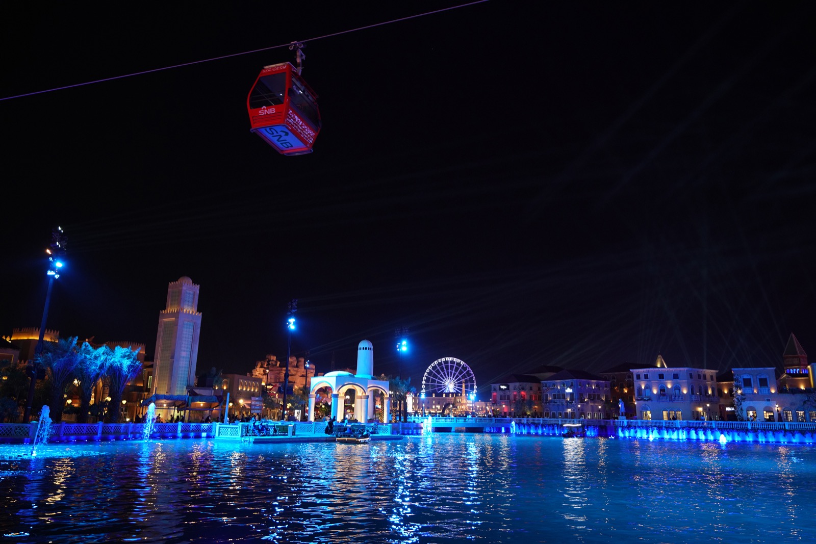 Boulevard Riyadh City illuminated at night with a cable car, Ferris wheel and blue-lit waterfront during Riyadh Season