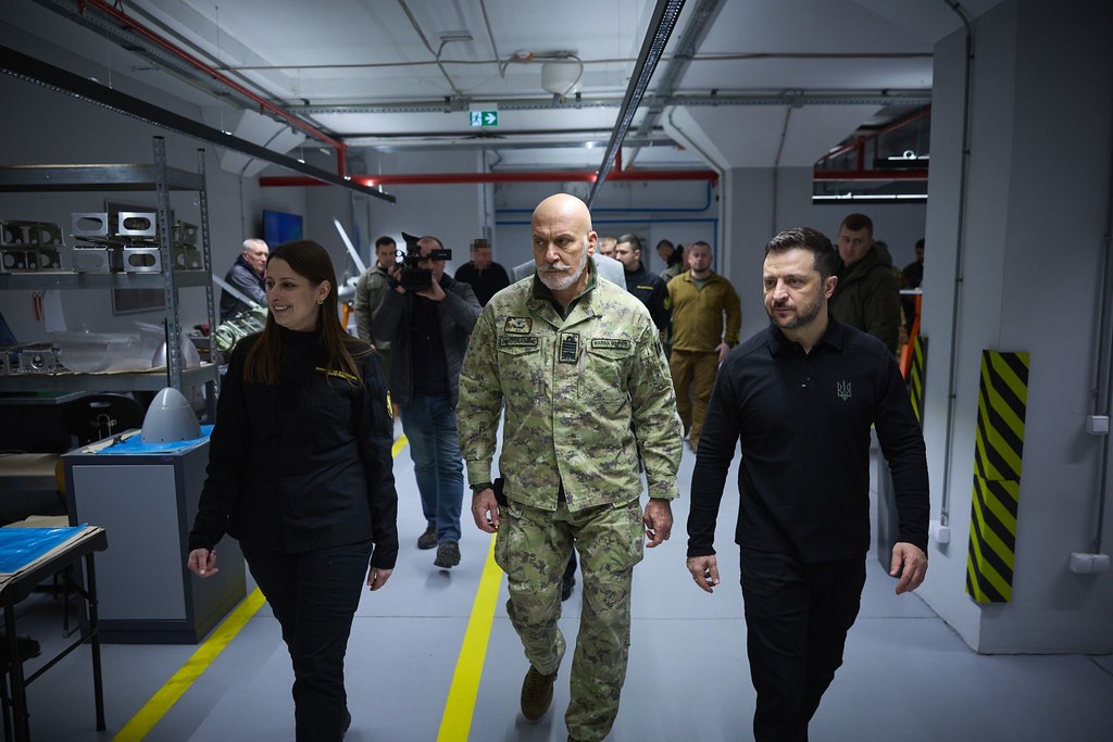 President Volodymyr Zelenskyy walks through a Ukrainian defense production facility alongside NATO Military Committee Chair Giuseppe Cavo Dragone, with drone and weapons hardware visible on assembly shelves