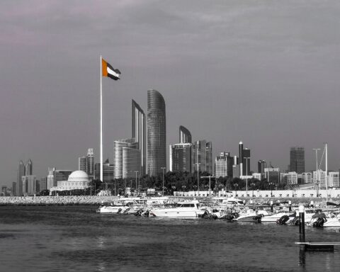 Abu Dhabi skyline with UAE national flag flying in the foreground, the Corniche waterfront and city towers behind, symbolising the UAE capital’s growing independence from Gulf institutional frameworks